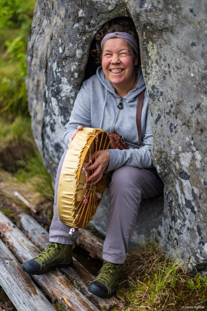 Henkilö istuu suuren kallion muodostamassa kapeassa kolossa ulkona luonnossa. Hän hymyilee leveästi ja pitelee koristeellista käsirumpua, jossa on vaaleanpunaiset nauhat. Päällä on harmaa huppari, vaaleat housut ja vihreät vaelluskengät. Maassa näkyy ruohoa ja puisia lankkuja.